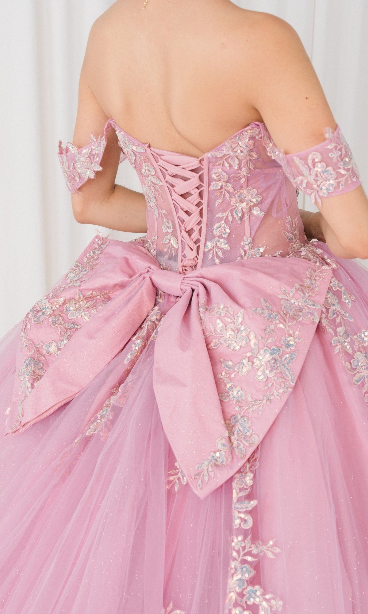 A person in the Quinceanera Dress 8405JQ by Cinderella Couture, an off-the-shoulder ball gown with pink fabric, silver floral embroidery and applique, a lace-up back, and a large waist bow.